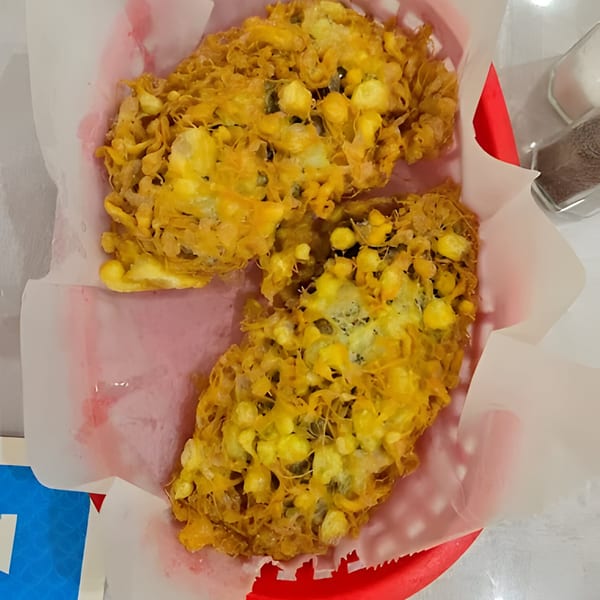 Two golden-brown corn fritters, crispy and irregular in shape, served in a red basket lined with white paper.